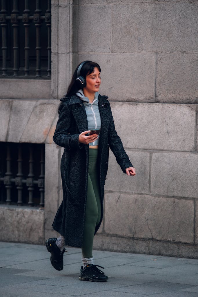 A woman walking on a city sidewalk in Madrid, wearing a coat and headphones.