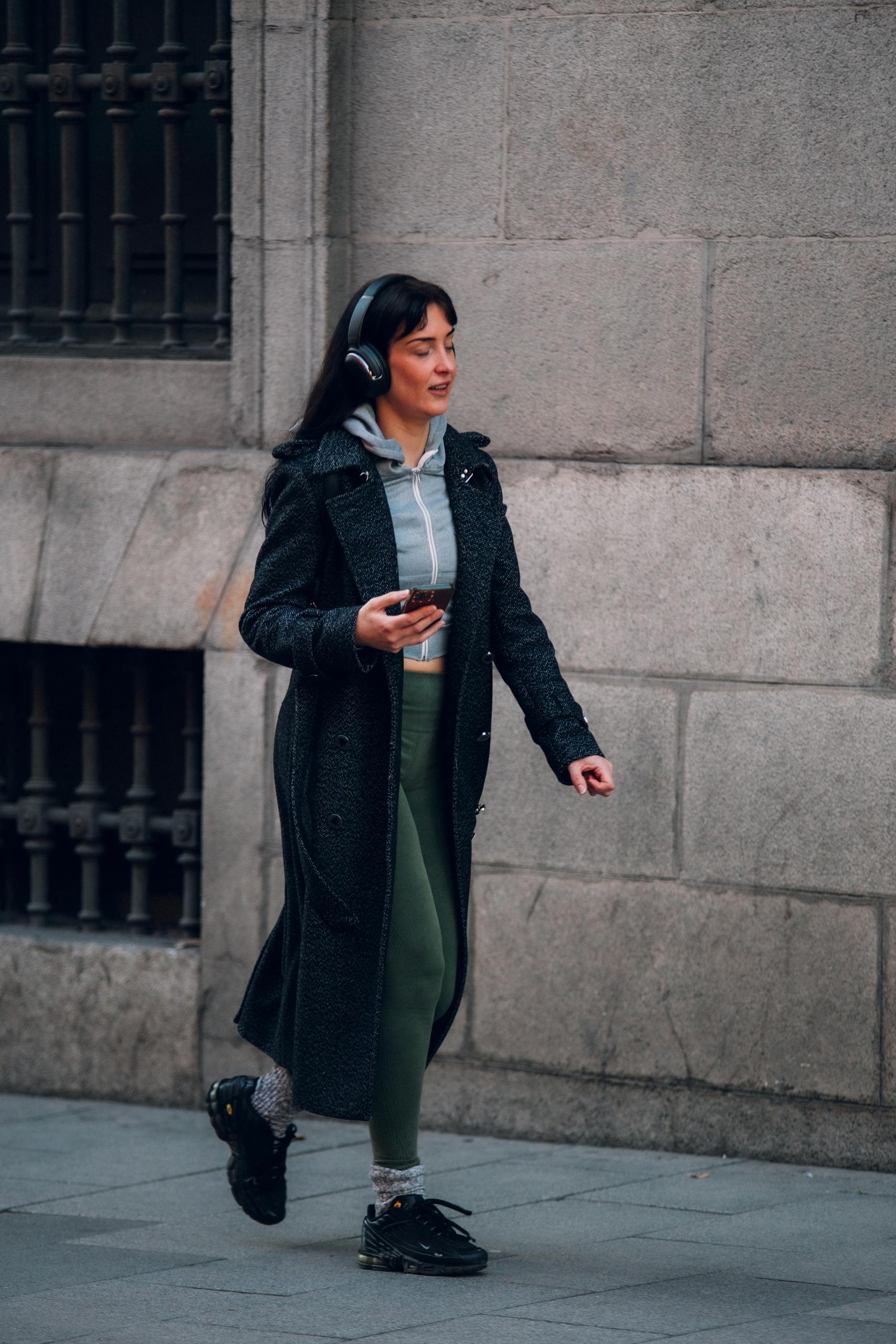 A woman walking on a city sidewalk in Madrid, wearing a coat and headphones.