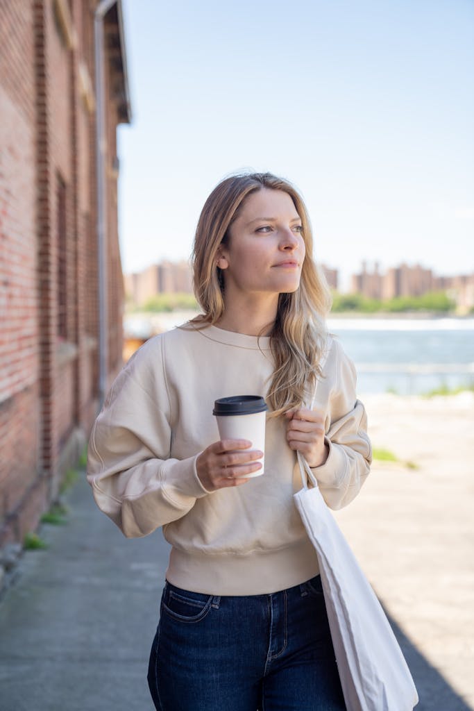 A woman walks along a riverside with a coffee cup and tote bag, enjoying a sunny day.