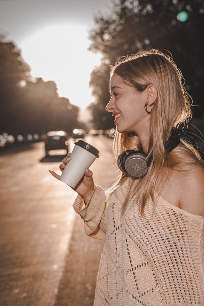 Smiling woman holding a cup in a sunlit city street with headphones around her neck.