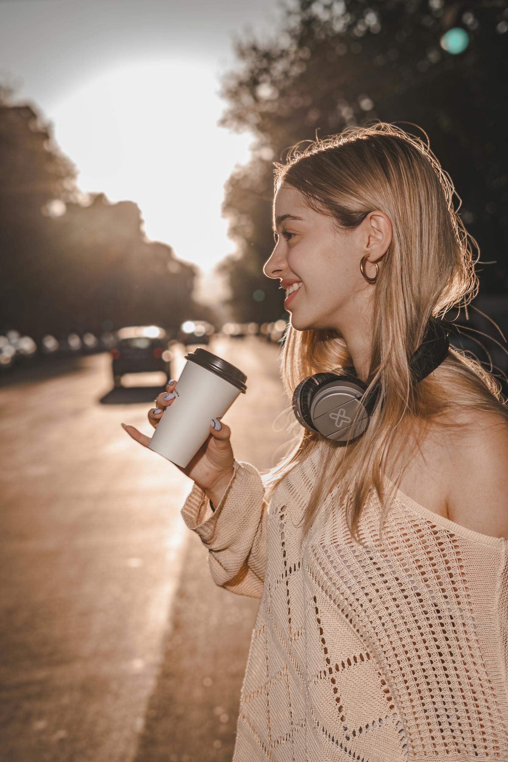 Smiling woman holding a cup in a sunlit city street with headphones around her neck.